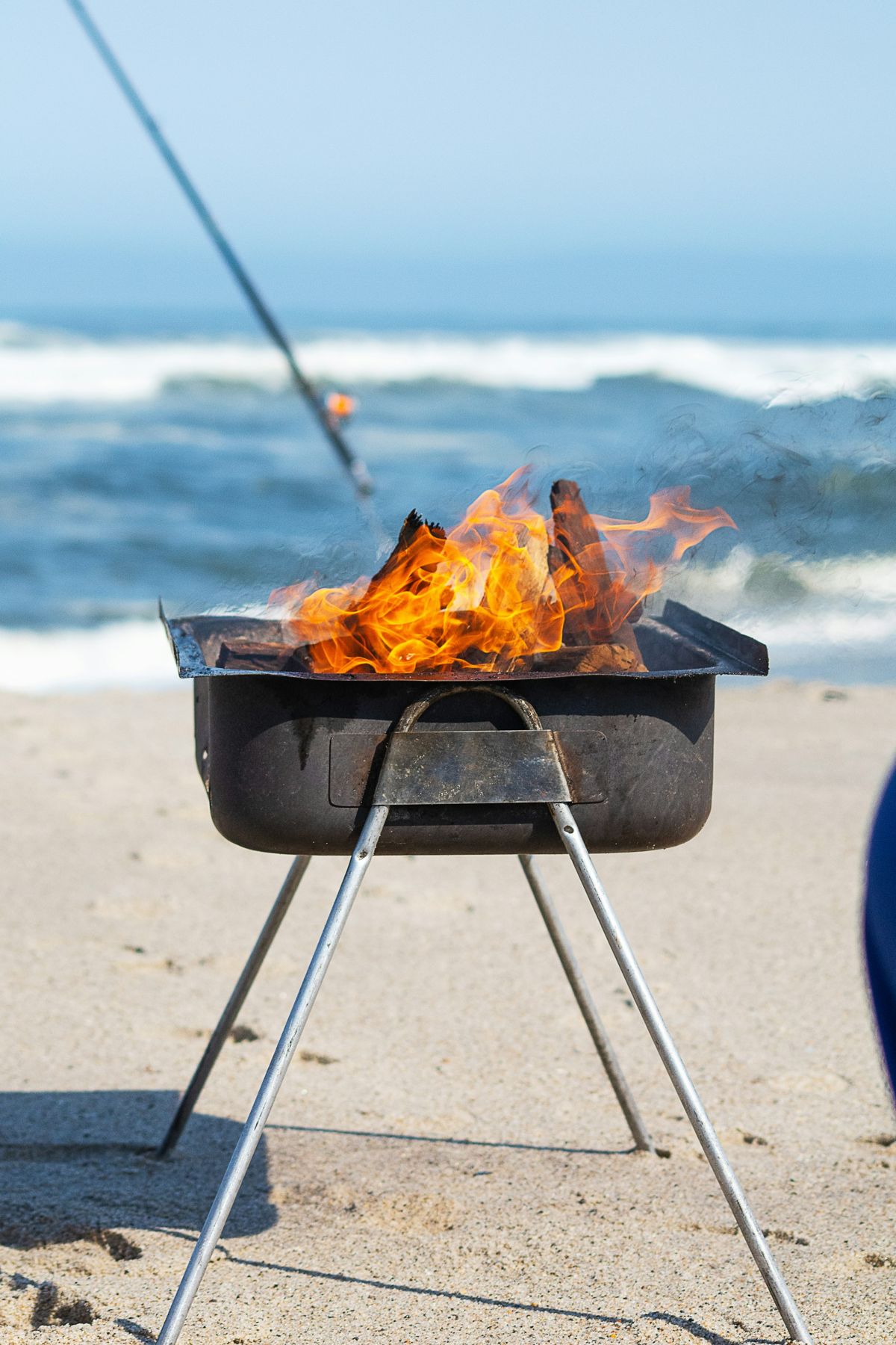 A braai with a fire going on a beach, a fishing rod beside it